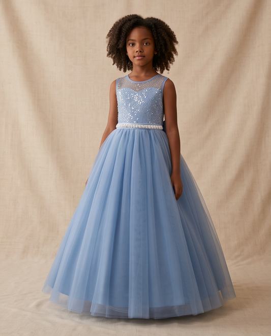 A young girl with curly dark hair stands wearing a sleeveless light blue ball gown with a sequined bodice and a full tulle skirt, posed against a neutral beige fabric backdrop.