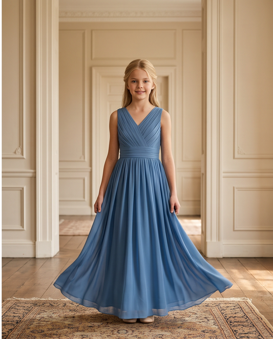 A young girl with blonde hair stands in a grand, elegant room wearing a sleeveless, V-neck blue maxi dress with a flowing skirt.