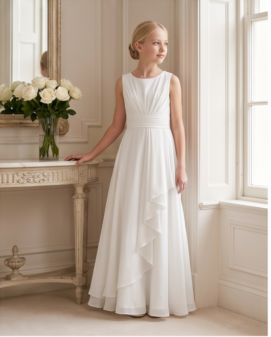 A young girl with blonde hair in a bun stands in a bright, elegant room wearing a sleeveless white A-line dress with a pleated bodice and a layered ruffle hem. She is posed next to a console table with a vase of white roses.