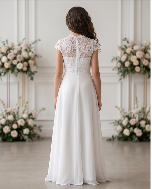 A woman stands with her back to the camera wearing a white wedding dress with a lace back and cap sleeves, photographed in a bright indoor setting with floral arrangements.