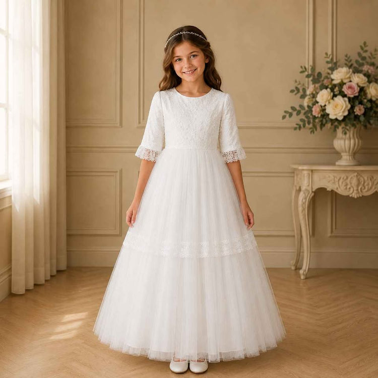 Young girl in a white lace dress standing in a room with beige walls and a table with flowers.