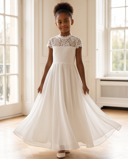 A young girl with her hair in a high bun stands in a bright room wearing a white A-line flower girl dress with a lace bodice and short cap sleeves. She is holding out the sides of her skirt to show its full flow.