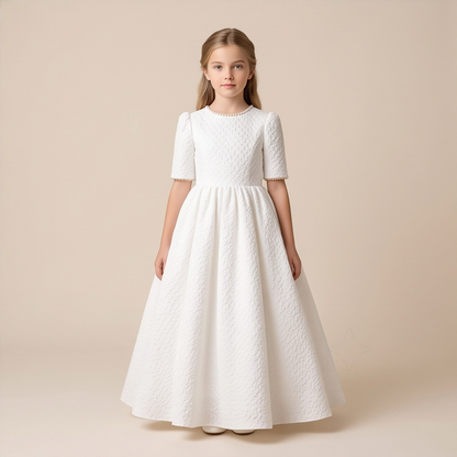A young girl stands against a plain beige studio background wearing a long white textured dress with short sleeves, a round neckline, and a full skirt.