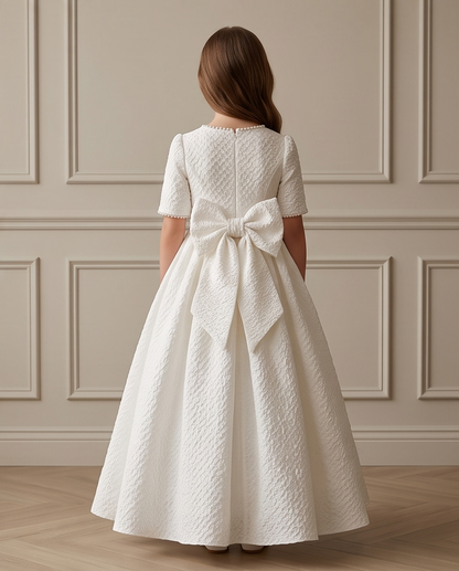 A young girl viewed from behind wearing a short-sleeved white first communion dress with a large bow at the back, standing in front of a beige paneled wall.