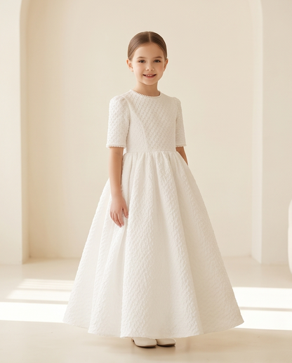 A young girl stands in a bright, minimalist cream-colored room wearing a formal white first communion dress with short textured sleeves and a full skirt.