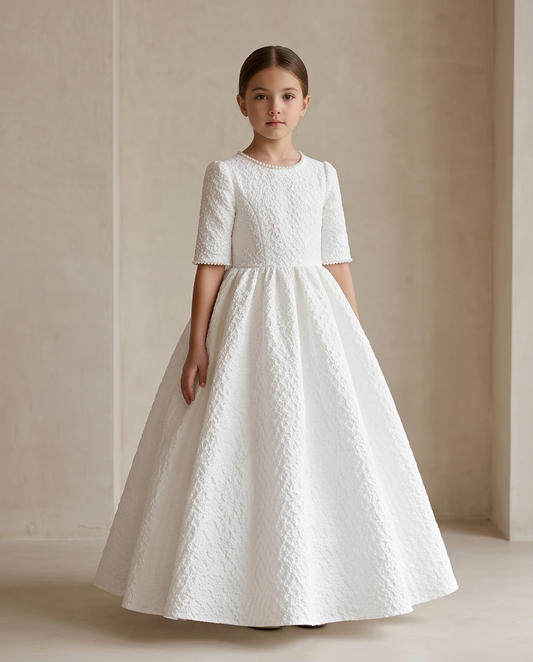 A young girl wearing a white textured A-line first communion dress with short sleeves, standing against a neutral beige wall.