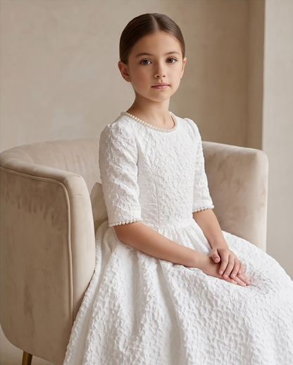 A young girl with dark hair sits on a beige velvet armchair wearing a white textured lace first communion dress with short sleeves and a pearl collar.