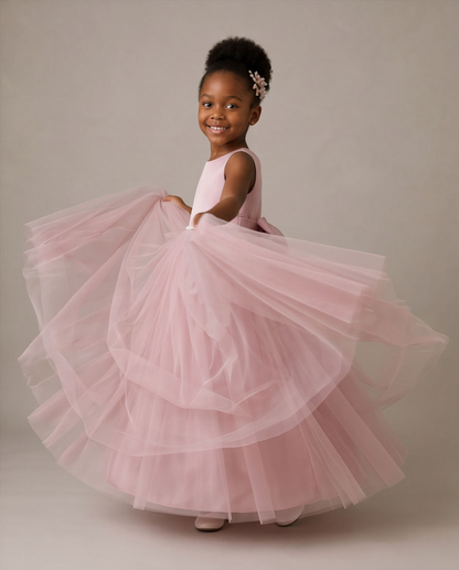 A young girl with her hair in a high bun wearing a light pink sleeveless dress with a voluminous, layered tulle skirt, posing and twirling to show off the dress against a neutral studio backdrop.