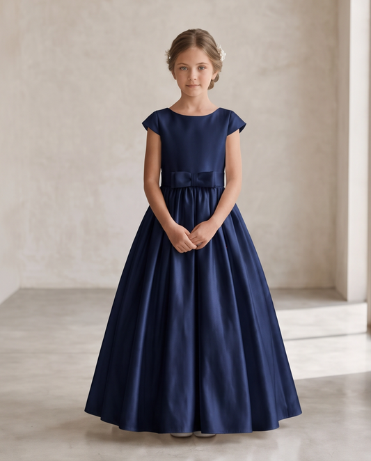 A young girl wearing a navy blue satin ball gown with cap sleeves and a matching waist bow, standing in a neutral studio setting.