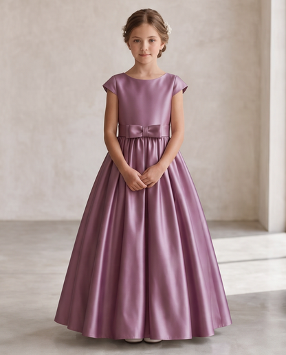 A young girl wearing a floor-length lavender satin dress with cap sleeves and a matching bow at the waist, standing against a neutral beige wall.