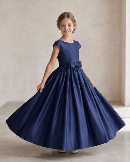 A young girl with light brown hair wearing a navy blue satin flower girl dress with a bow at the waist, standing in a studio with a textured beige wall background.
