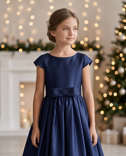 A young girl with her hair in a low bun wears a navy blue satin flower girl dress with cap sleeves and a matching waist bow. She is standing in a festive indoor setting with a blurred Christmas tree and warm bokeh lights in the background.