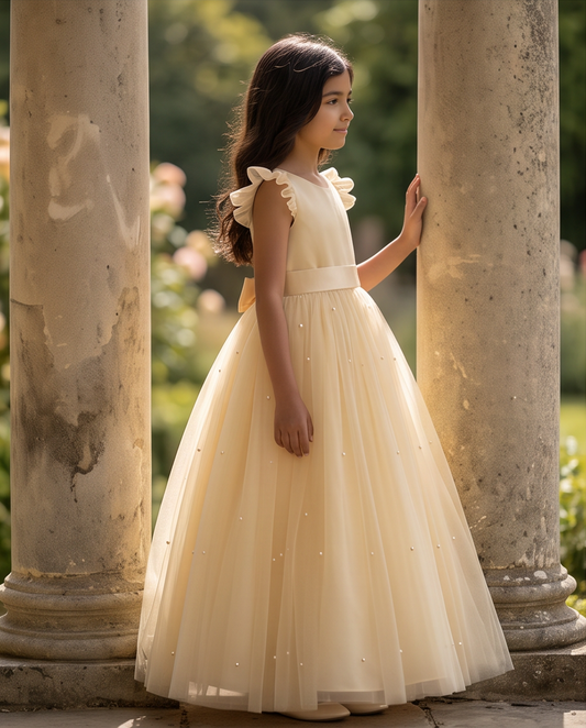 A young girl wearing a cream tulle ball gown with ruffled shoulder straps and a matching waistband, standing between two stone columns in a sunlit garden setting.