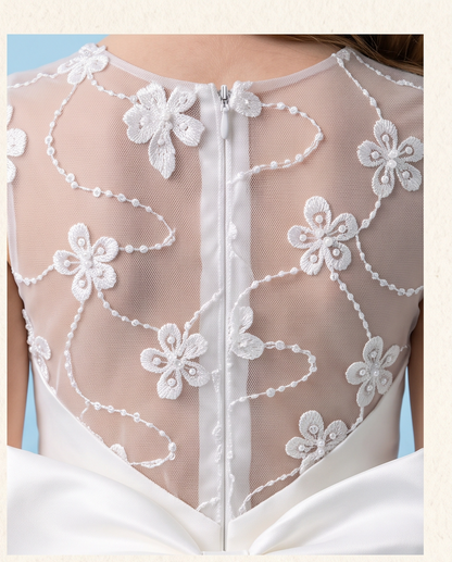Close-up of the back of a bridal gown showing a sheer ivory mesh back panel with white floral applique, bead trim, and a central zipper, with a white satin bow at the waist.