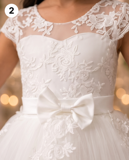 White lace and tulle dress with a large satin bow at the waist, shown on a model against a warm blurred bokeh background. A small white circle with the number 2 appears in the top-left corner.