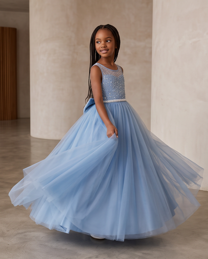 A young girl with long dark braids is wearing a sleeveless light blue tulle dress with a beaded embellished bodice and a full flowing skirt, posing in a spacious indoor setting with beige columns.