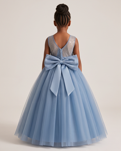 A young girl viewed from behind wearing a sleeveless light blue tulle ball gown with a large bow at the back and a sparkly silver sequined V-neck bodice, photographed against a plain light beige studio background.