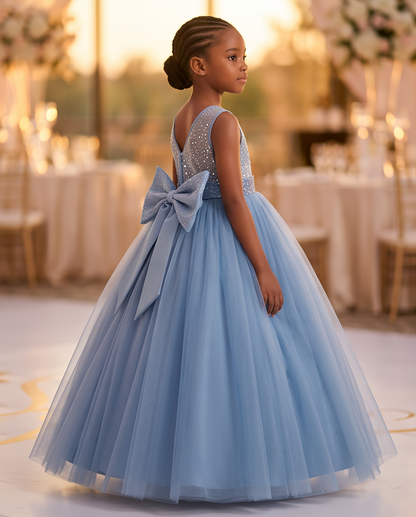 A young girl stands in profile wearing a formal light blue ball gown with a beaded bodice, a large bow at the back, and a full tulle skirt in an indoor event setting at golden hour.