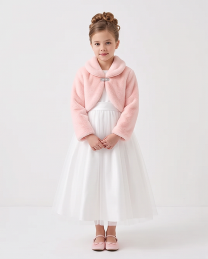 A young girl with her hair in two buns stands against a plain white studio background, wearing a light pink fuzzy cropped jacket with a silver button over a white tulle dress and pink shoes.