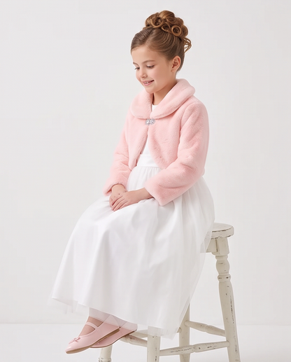A young girl with her hair in rolls sits on a distressed white stool, wearing a light pink faux-fur collar jacket over a white dress and pink flats.