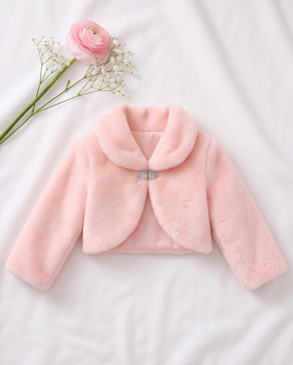 A light pink faux fur jacket for toddlers, featuring a wide collar and a decorative silver bow at the center front, laid flat on a white fabric background with a pink rose and baby's breath flower as props.