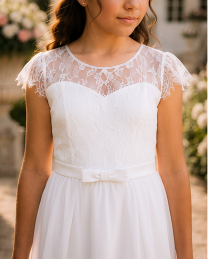 A girl wearing a white dress with a sheer lace yoke, flutter sleeves, and a matching satin waist bow, photographed outdoors with a softly blurred floral background.