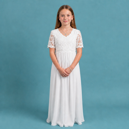 A young girl with long brown hair stands against a solid teal-blue studio background wearing a long white dress with a lace bodice and lace short sleeves.