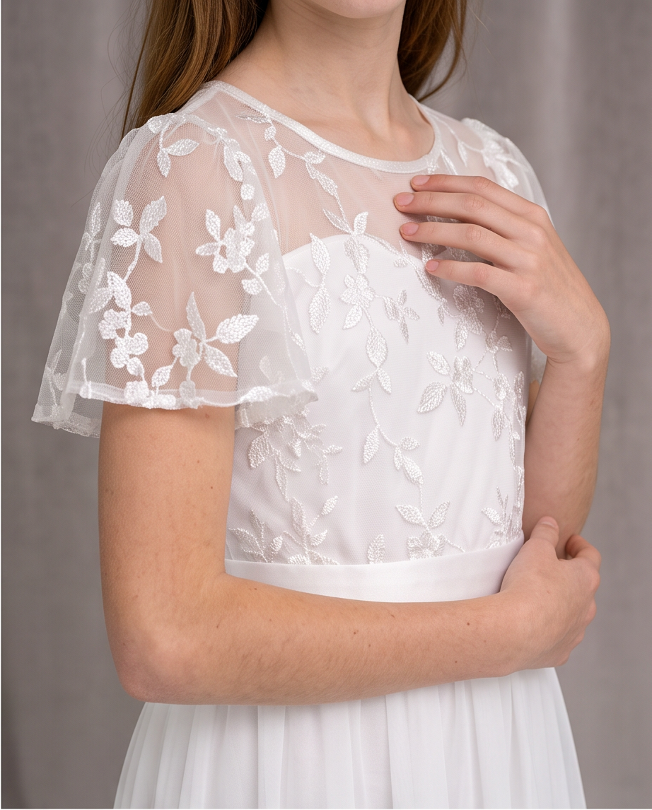 A close-up of a model wearing a white lace and mesh short-sleeve top with floral embroidery, paired with a white skirt, against a neutral gray background.