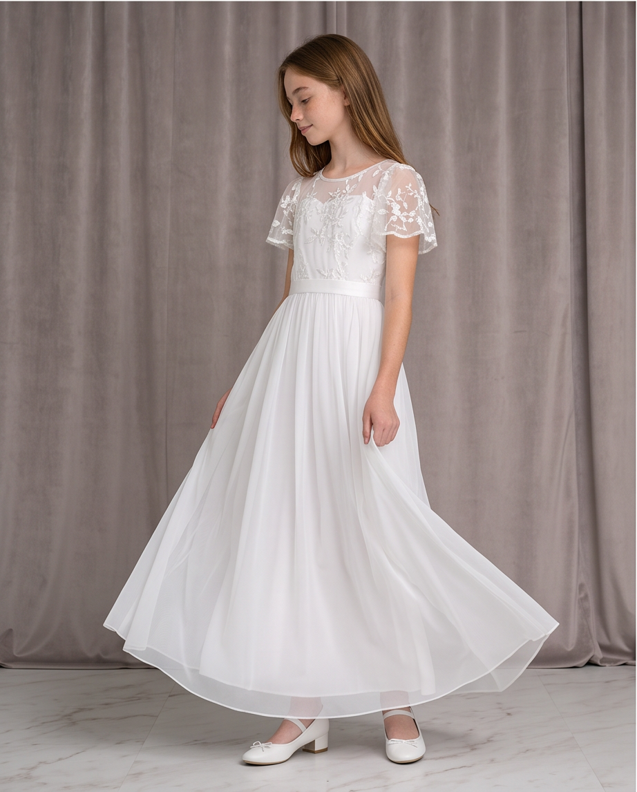 A young girl wearing a long white first communion dress with lace short sleeves and a flowing chiffon skirt, standing in front of a gray curtain backdrop.