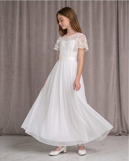 A young girl wearing a long white first communion dress with lace short sleeves and a flowing chiffon skirt, standing in front of a gray curtain backdrop.
