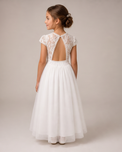 A young girl viewed from the back wearing a white formal dress with a lace open-back bodice, short cap sleeves, and a full tulle skirt, photographed against a plain warm beige studio background.