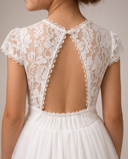Close-up back view of a white lace dress with a sheer floral lace open back, short puff sleeves, and a gathered white skirt against a light neutral background.