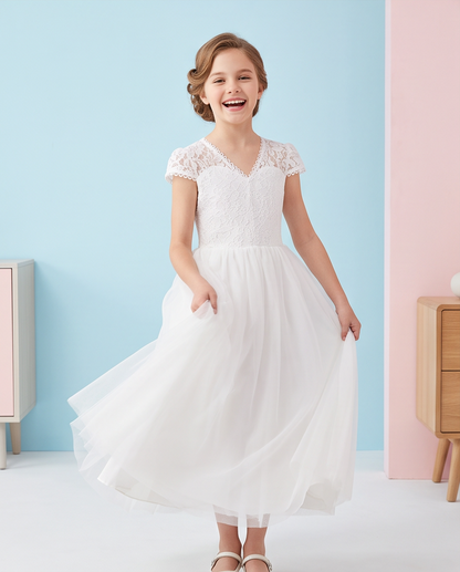 A young girl with styled brown hair is smiling and twirling in a white lace and tulle flower girl dress, standing against a pastel blue and pink studio wall.