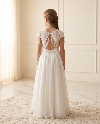 A young girl viewed from behind wearing a white A-line dress with a lace open back and tassel fringe, standing on a cream rug in a bright room.