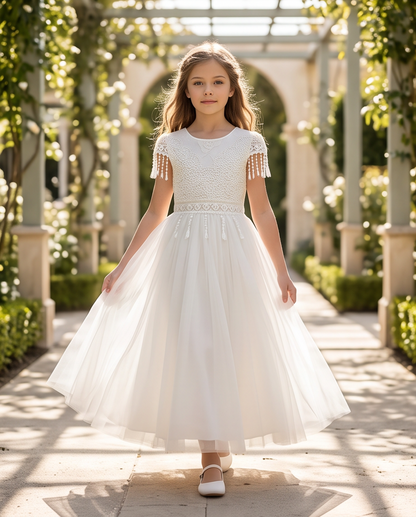 A young girl wearing a white first communion dress with short cap sleeves, a textured lace bodice, and a full tulle skirt, walking down a sunlit garden path lined with greenery.