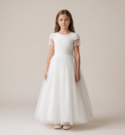 A young girl stands against a warm beige studio background wearing a white formal dress with a textured lace bodice, short cap sleeves with tassel fringe, and a full tulle skirt. Text at the bottom reads, 'FIND YOUR PERFECT LOOK Shop The Collection at [Website/Brand Name]'.