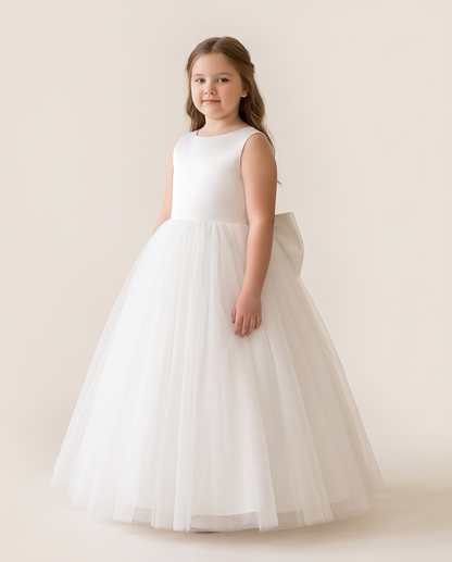 A young girl with light brown hair stands wearing a sleeveless white formal dress with a fitted bodice and a very full tulle skirt. She is photographed against a plain, warm off-white studio background.