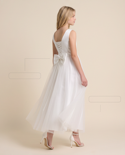 A girl modeling a sleeveless white wedding dress from the back, featuring a lace-up open back, a large bow at the waist, and a flowing tulle skirt, against a solid beige studio background.