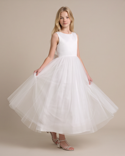 A young girl wearing a sleeveless white dress with a fitted bodice and a full tulle skirt, posing with her hands on the sides of the skirt against a solid beige studio background.