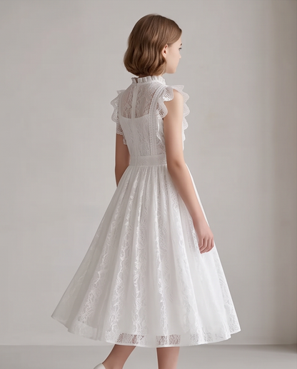 A girl stands on a sunlit garden path wearing a white lace midi dress with ruffled cap sleeves, a high neckline, and a full skirt. 
