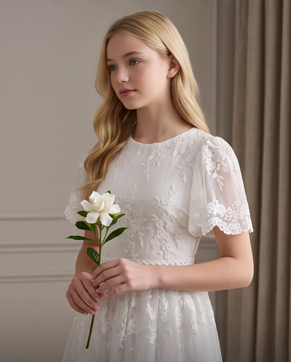 A young blonde woman wearing a white lace and tulle dress, holding a white flower, posed against a neutral indoor background with text overlay at the bottom.