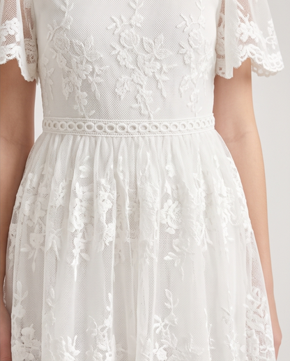 Close-up of a white lace dress with floral embroidery, short flutter sleeves, and a gathered tulle skirt, shown on a model against a light neutral background.