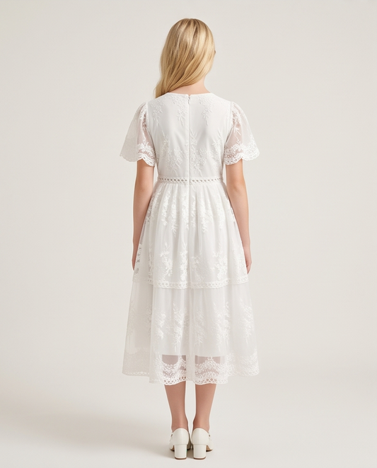 A girl viewed from the back wearing a white midi dress with short flutter sleeves, lace trim, and a tiered skirt, photographed against a plain light beige studio background.