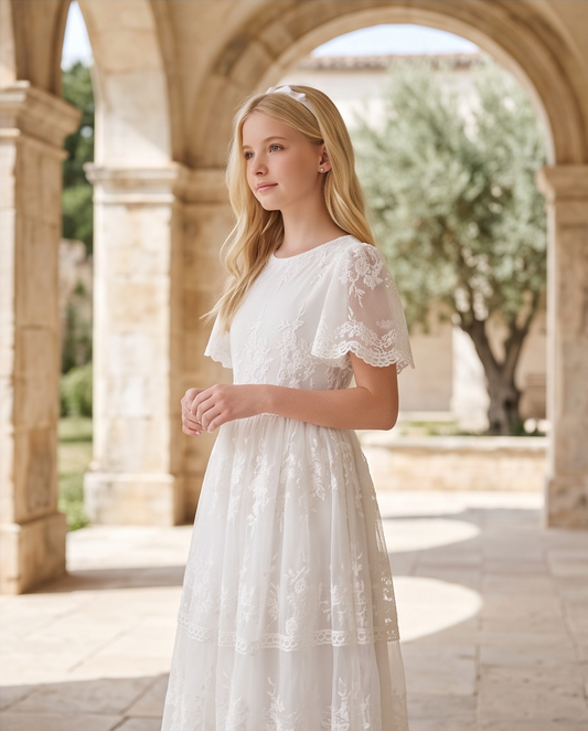 A young girl with long blonde hair stands in a stone colonnade wearing a white lace and tulle dress. The dress has short flutter sleeves with lace trim, a round neckline, and a tiered skirt with embroidered floral details.