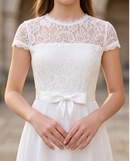 A woman wearing a white lace and satin dress with short cap sleeves, a lace bodice, a satin waistband with a bow, and a flowing skirt. The photo is cropped from the lower face to the hips against a softly blurred outdoor background.
