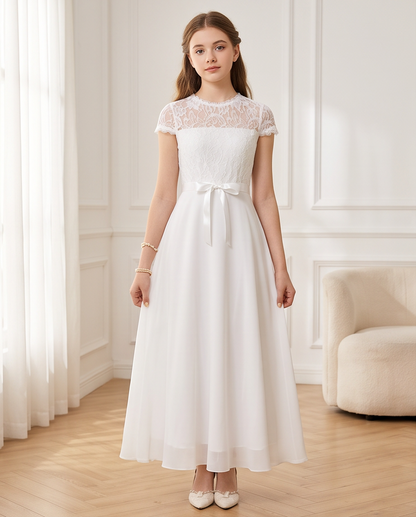 A young woman stands indoors wearing a white lace and satin midi dress with a tied waist and cream pointed flats. The room has white paneled walls, sheer curtains, and a light upholstered chair.