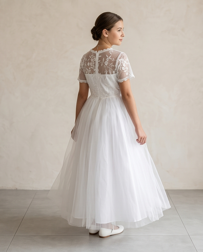 A girl stands on a sunlit garden path wearing a white lace midi dress with ruffled cap sleeves, a high neckline, and a full skirt. 