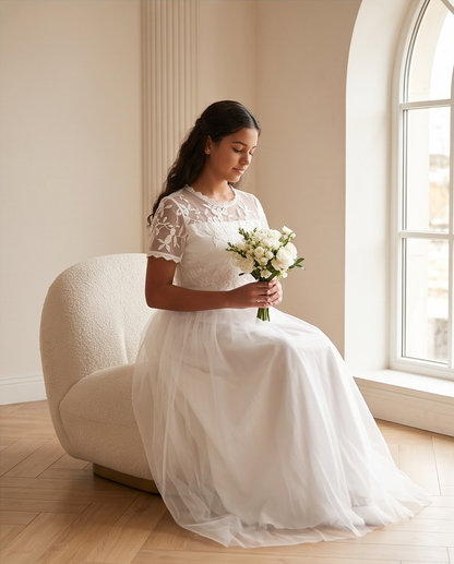 A girl sits on a sunlit garden path wearing a white lace midi dress with ruffled cap sleeves, a high neckline, and a full skirt. 