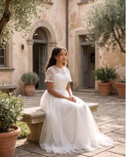 A woman wearing a white lace and tulle wedding dress sits on a stone bench in a sunlit courtyard with potted plants and a beige building in the background.