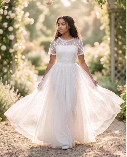 A girl stands on a sunlit garden path wearing a white lace midi dress with ruffled cap sleeves, a high neckline, and a full skirt. 